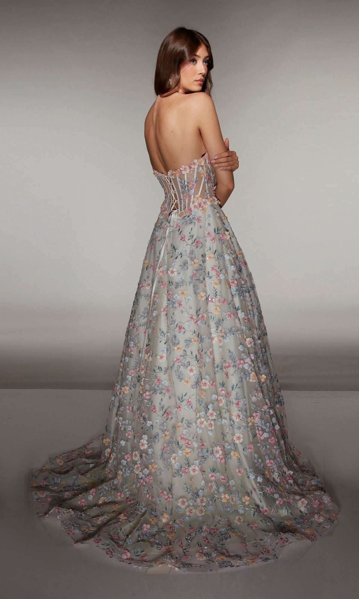 A woman stands facing away, wearing the Alyce 61986 long prom dress—a strapless A-line gown with a sheer corset bodice decorated with 3D floral appliques.